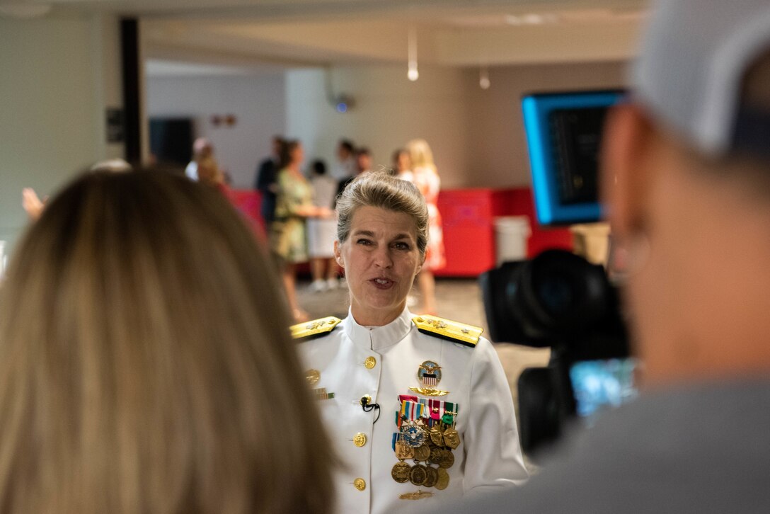 A woman in Navy dress whites (skirt version) with gold trim and many medals and ribbons chats with two people.