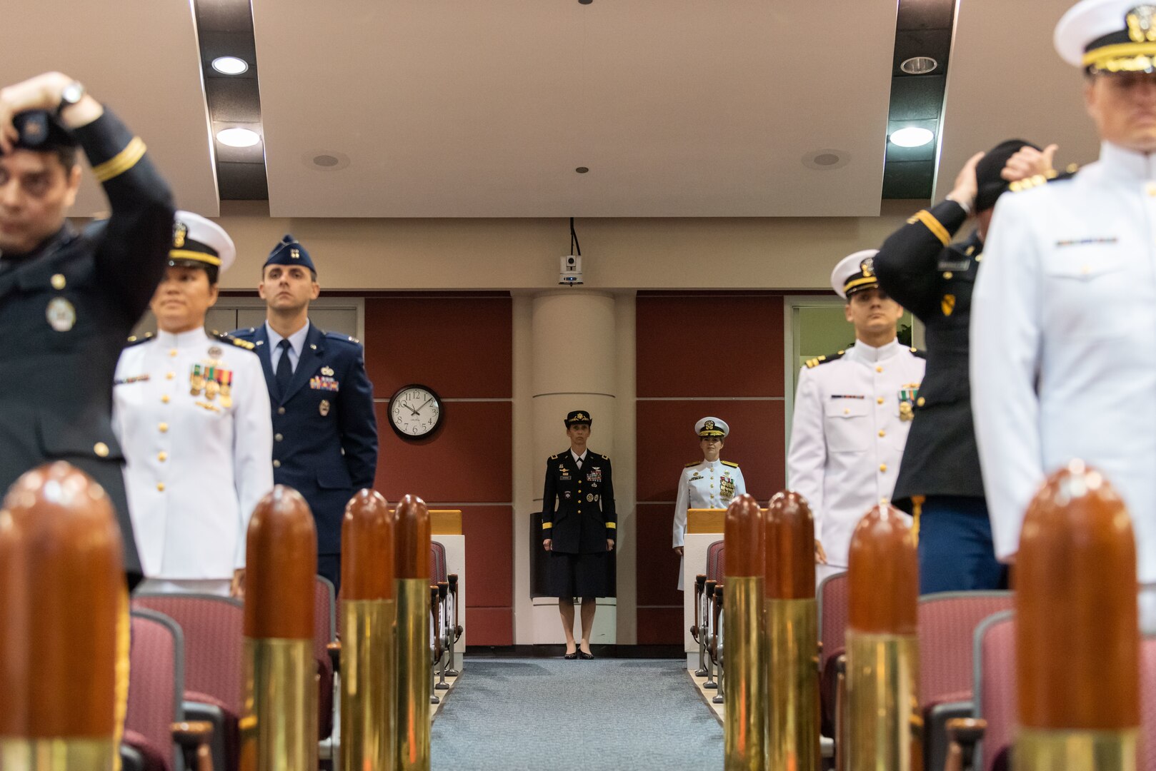 A woman in black stands at the back of a auditorium with a woman in white. Military members in Navy and Army dress uniforms flank the women in the back with reproductions of bullets on either side. The woman in black is in Army dress blues. The woman in white is in Navy dress whites. Two Army men are straightening their caps.