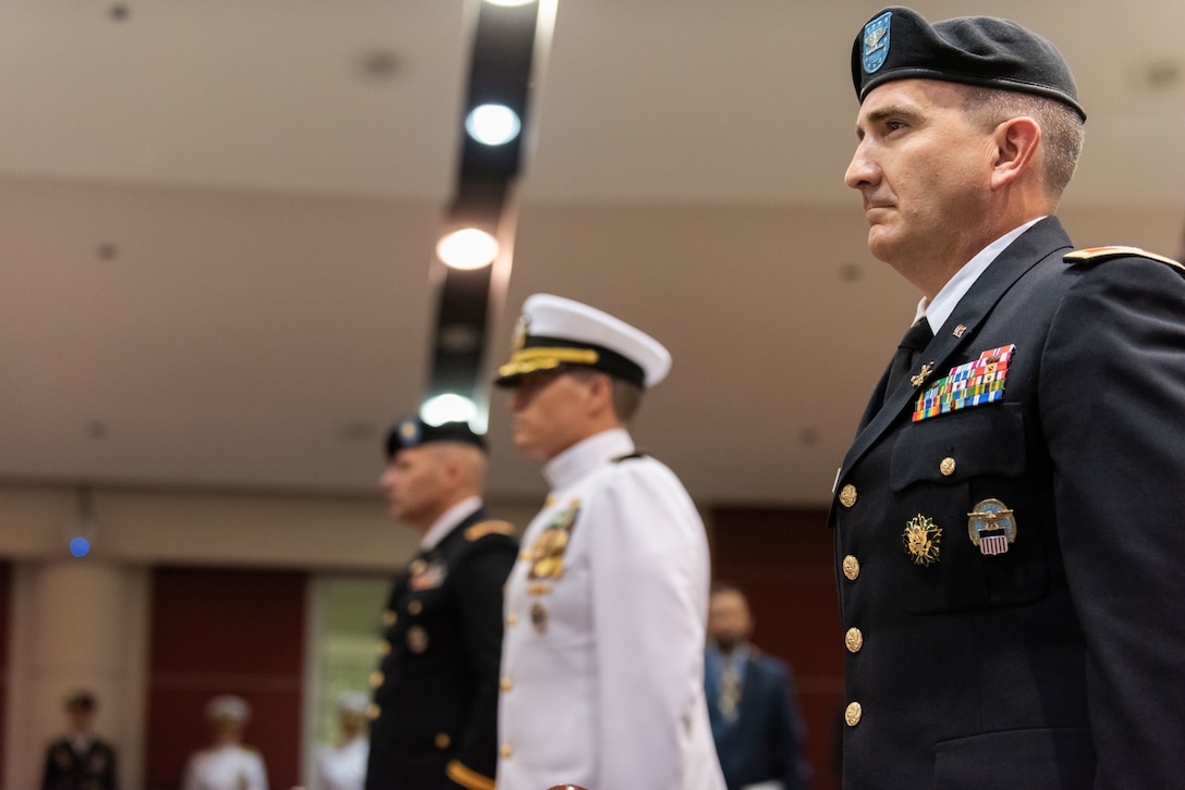 Three men, two in Army dress uniforms which are black in color with gold trim and one in the middle wearing Navy dress whites which is a pure white uniform with gold trim. They all have their hats on.