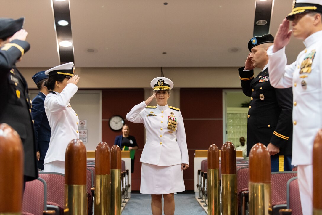 A woman in Navy dress whites (skirt version) with gold trim and many medals and ribbons salutes as she walks into a auditorium. She is flanked by large bullet reproductions and military members from the Air Force, Navy and Army acting as her Sideboys.