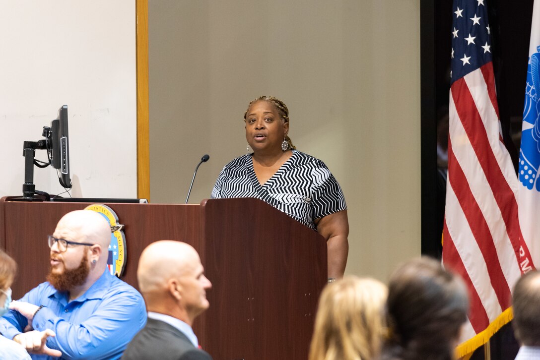 A heavyset woman with blond braids and a black and white patterned blouse sings at a podium in an auditorium.