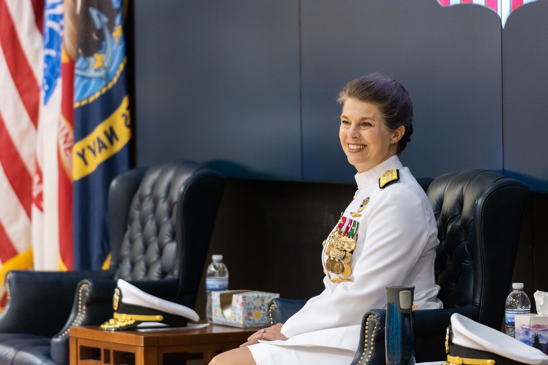 A woman with braided hair in a bun is seated on stage with her hands in her lap. She wears a pure white Navy uniform with gold trim and many medals and badges. She is smiling.