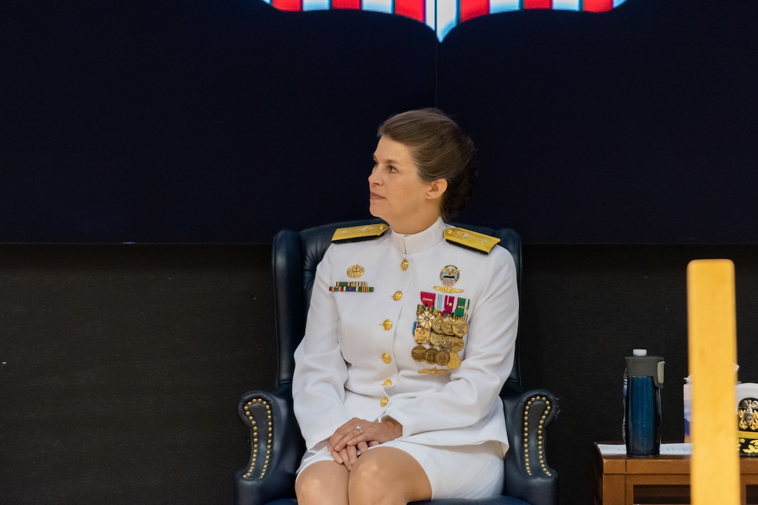 A woman with braided hair in a bun is seated on stage with her hands in her lap. She wears a pure white Navy uniform with gold trim and many medals and badges.