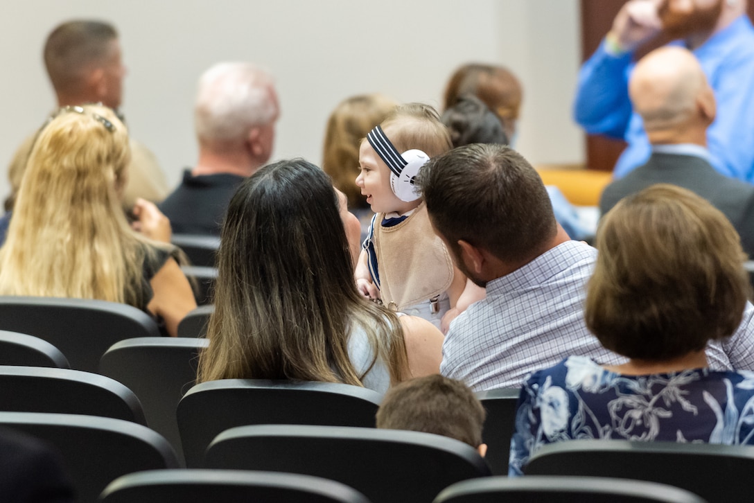 A shot of the backs of heads of several audience members. A couple in the middle are holding a baby up. The baby wears hearing protection.