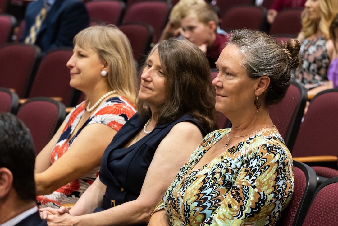 Three women in dresses are seated in an auditorium. One has graying hair, one has black hair and one has blond hair. They are all smiling.