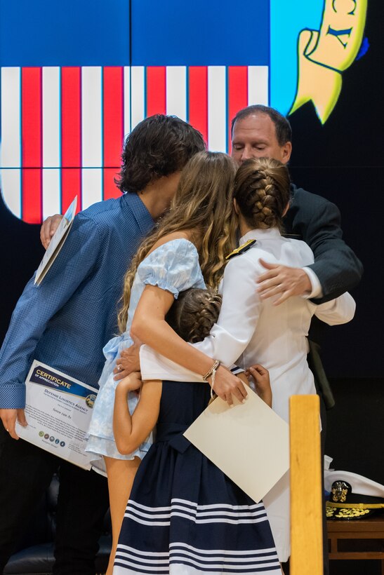 A woman with braided hair in a bun and dressed in Navy whites hugs her two adult children and her younger daughter and husband on stage after giving them family appreciation certificates. The DLA logo is projected on screen in the background.