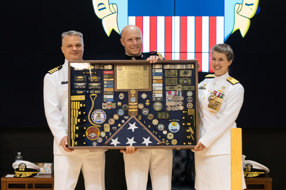 A woman with braided hair in a bun helps hold a large shadowbox with all sorts of medals, coins, badges, a folded flag, ranks etc. She wears a pure white Navy uniform with gold trim and many medals and badges. She is helped by her aide in a Marine Corps uniform and a man in Navy dress whites. They are standing on stage with the DLA logo in the background projected on a screen.