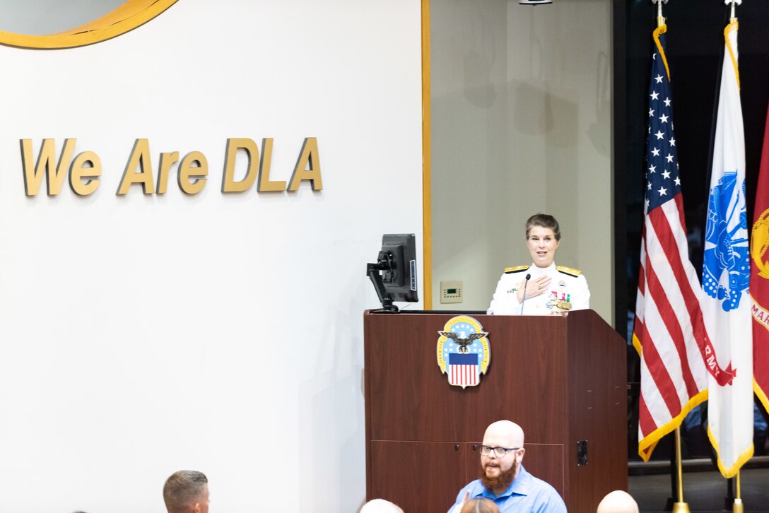 A woman with braided hair in a bun stands at a podium with a the US Flag and military flags in the background. She wears a pure white Navy uniform with gold trim and many medals and badges.