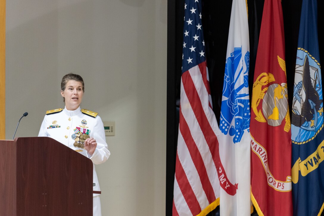 A woman with braided hair in a bun stands at a podium with a the US Flag and military flags in the background. She wears a pure white Navy uniform with gold trim and many medals and badges. She is lifting her hand in emphasis.