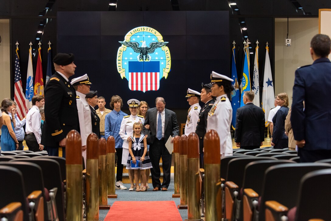 A group of people surrounding a woman in Navy dress whites prepare to walk down the aisle in a auditorium. The Navy uniform she wears is pure white. White dress coat with gold trim and gold epilets and white skirt with white shoes. She has two hands on her daughter in a blue and white dress and is flanked by her husband in a grey suit with blue tie. Behind her is her adult daughter and son in a blue dress shirt.