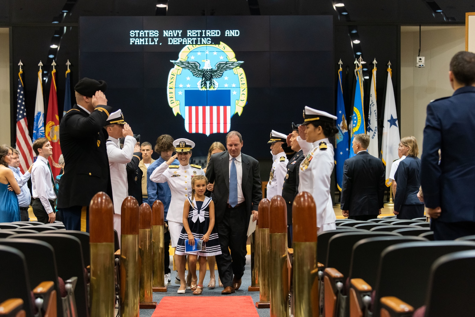 A woman in Navy dress whites is surrounded by her young daughter in a blue and white dress, a man in a dark suit with blue tie, a young adult male and female. They are walking down the aisle in an auditorium. She is saluting her Sideboys which is a group of military members flanking her on her right and left.