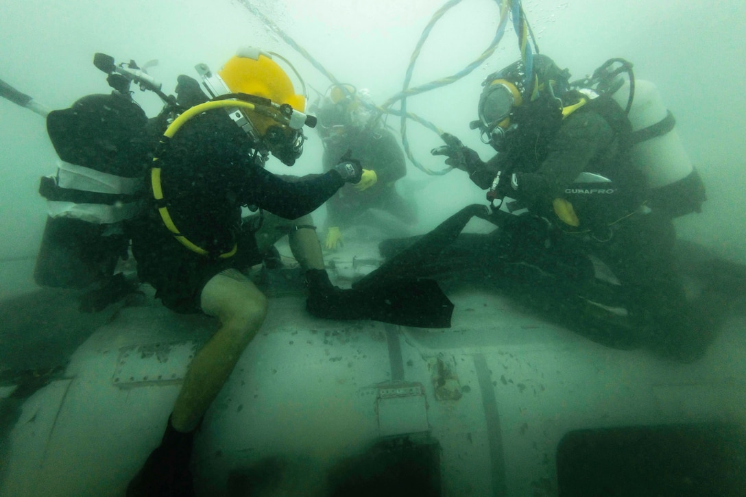 U.S. and Dutch naval forces wearing diving gear communicate while sitting on a piece of equipment underwater.