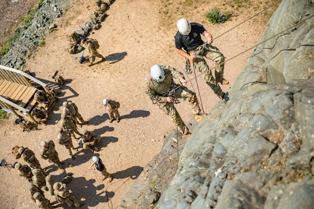 Two soldiers rappel down a rock toward a group on the ground.