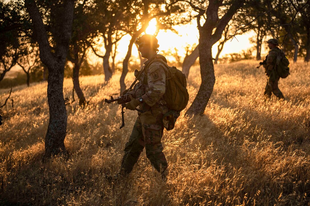 Soldiers walk through a wooded area as the sun shines behind.
