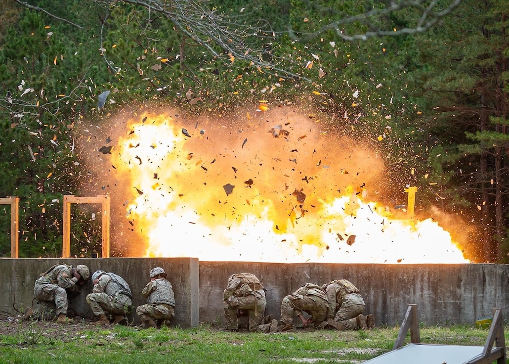 Soldiers take cover behind a low wall as explosion occurs.