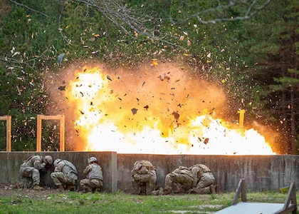 Soldiers take cover behind a low wall as explosion occurs.