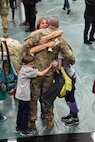 Soldier faces away from camera while his family simultaneously hugs him.