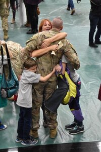 Soldier faces away from camera while his family simultaneously hugs him.
