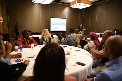 Groups of people sitting at tables receiving instruction.
