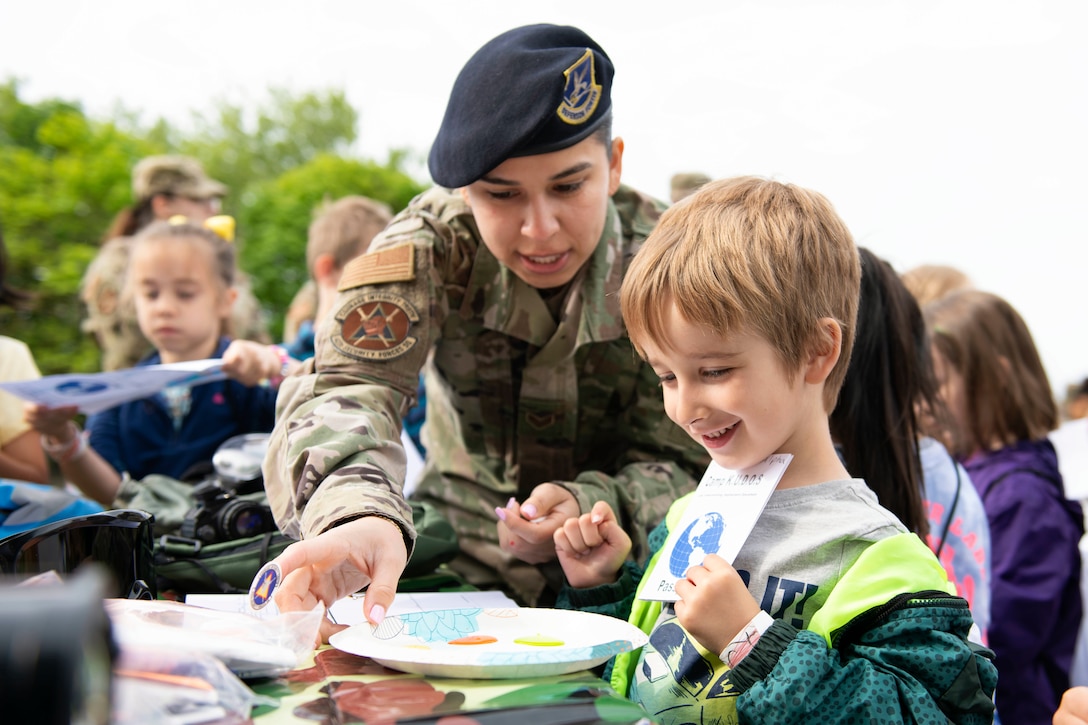 An airman gives a student sitting at a table with other students a plate with paint on it.