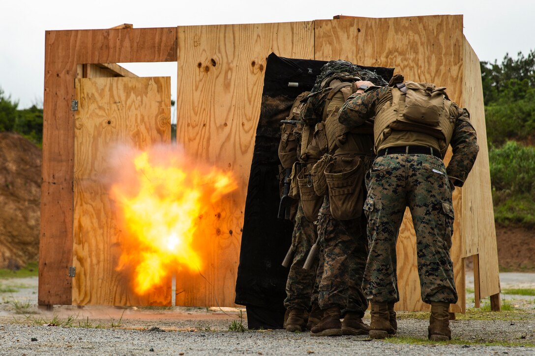 Marines stand behind a shield as an explosive detonates in a building made of plywood during training.