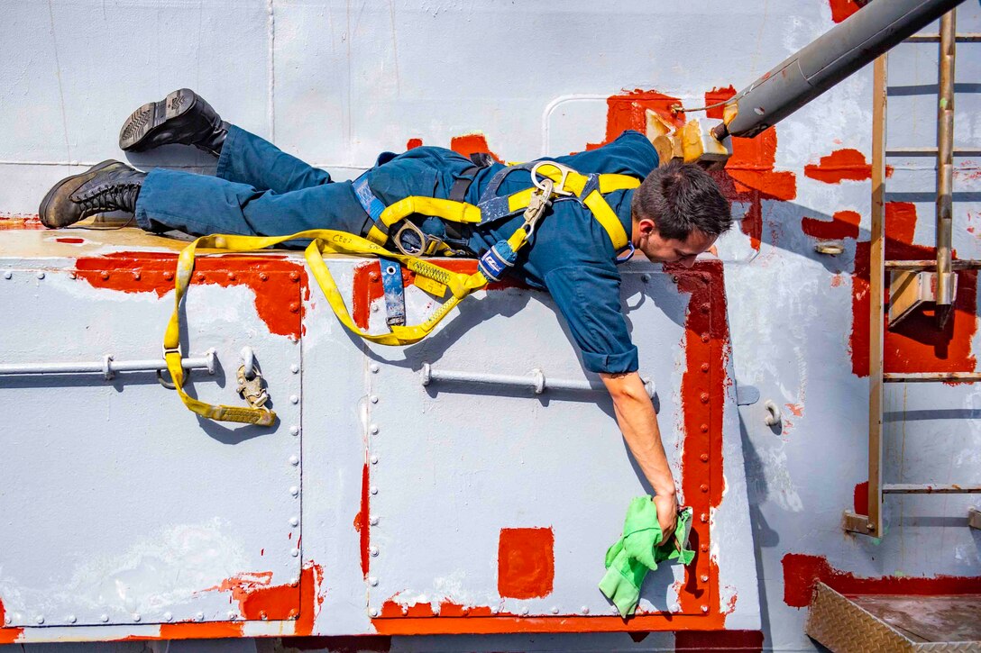 A sailor uses a rag to wipe a bulkhead on a ship.