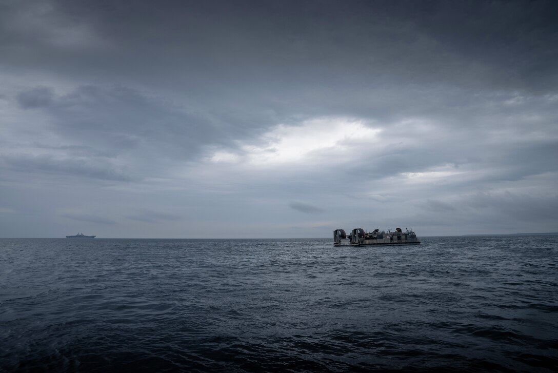 A landing craft travels away from a ship in the distance amid gray skies.
