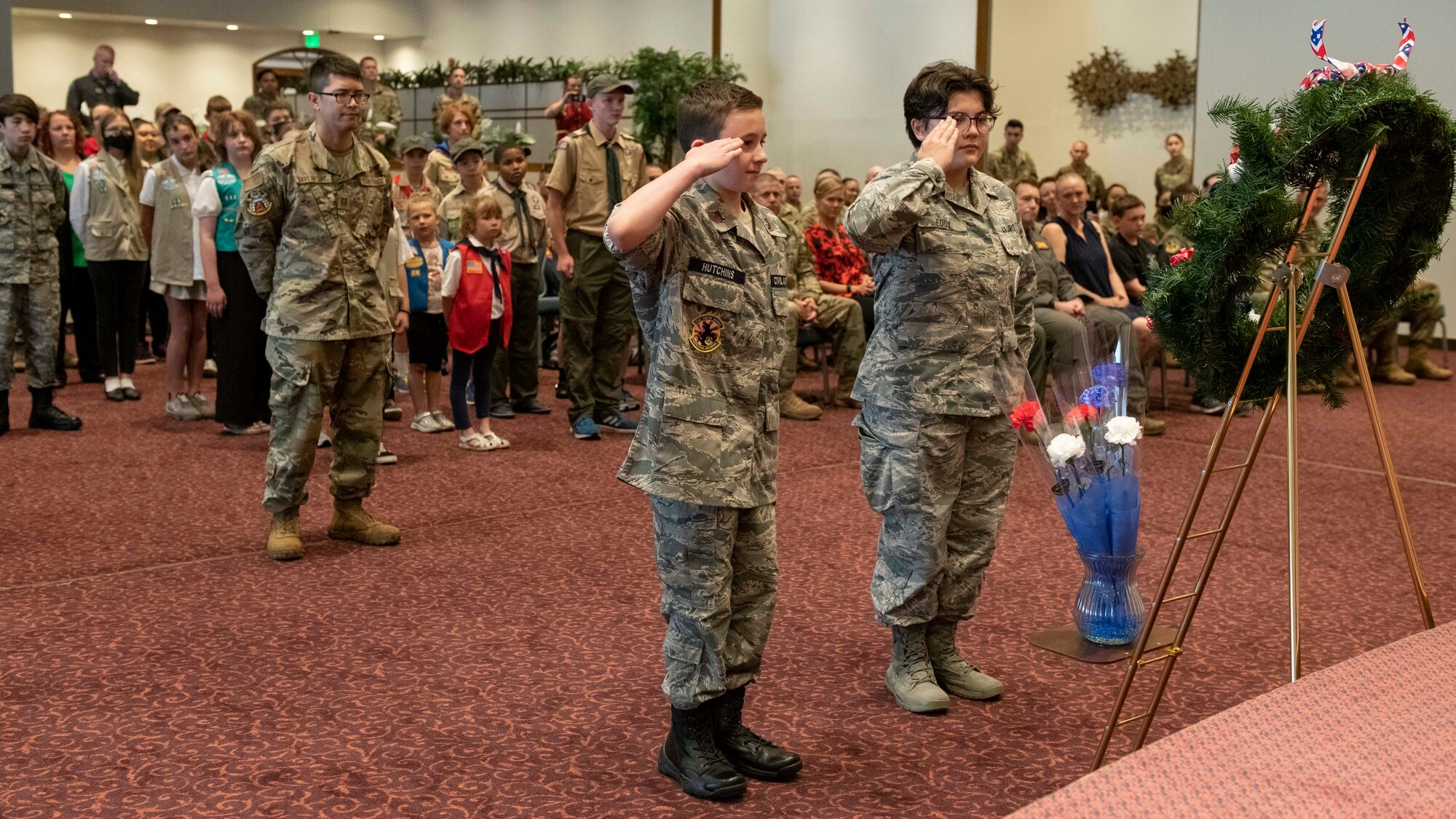 Two teenagers in military uniform salute a wreath in front of a stage.