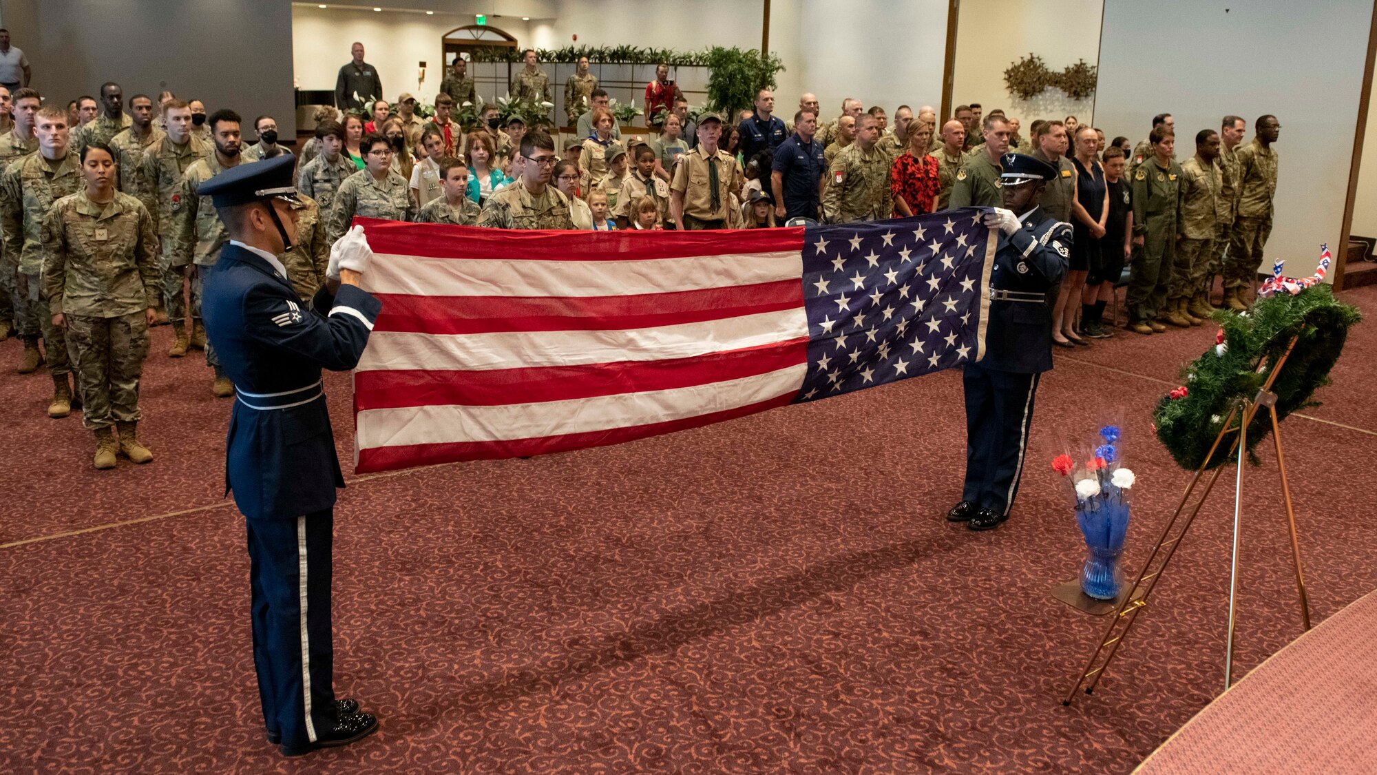 Two ornately dressed honor guards fold a large U.S. flag between them in front of a standing crowd.