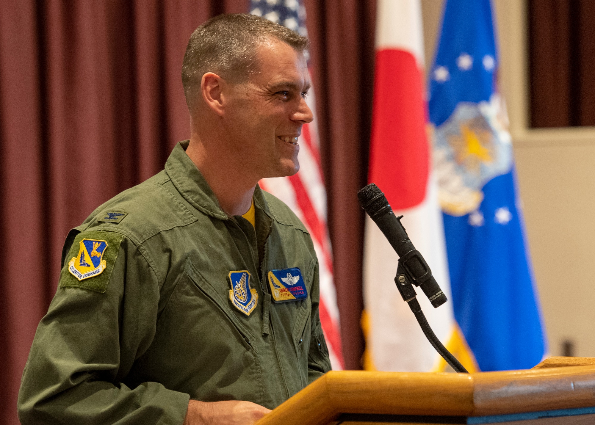 A male colonel in a flight suit smiles from a podium