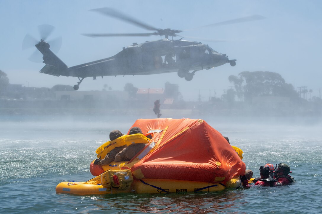A sailor hangs below a helicopter as Marines float in the water and sit on a raft.