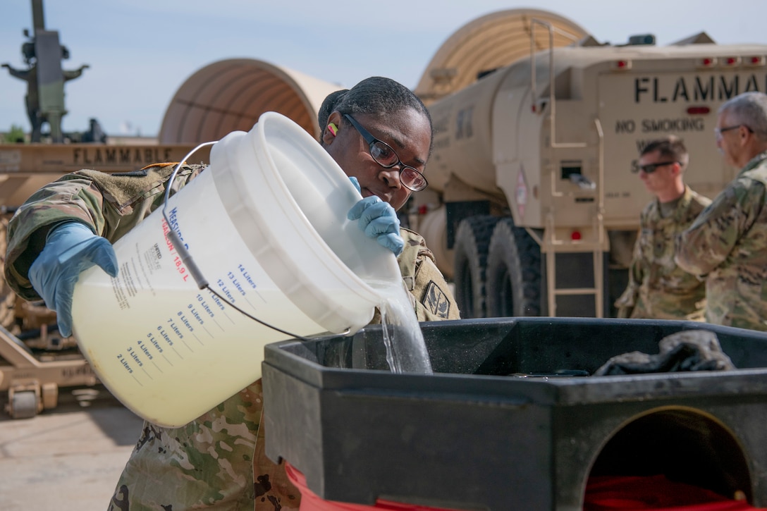 An airman pours a bucket of liquid into a large container.