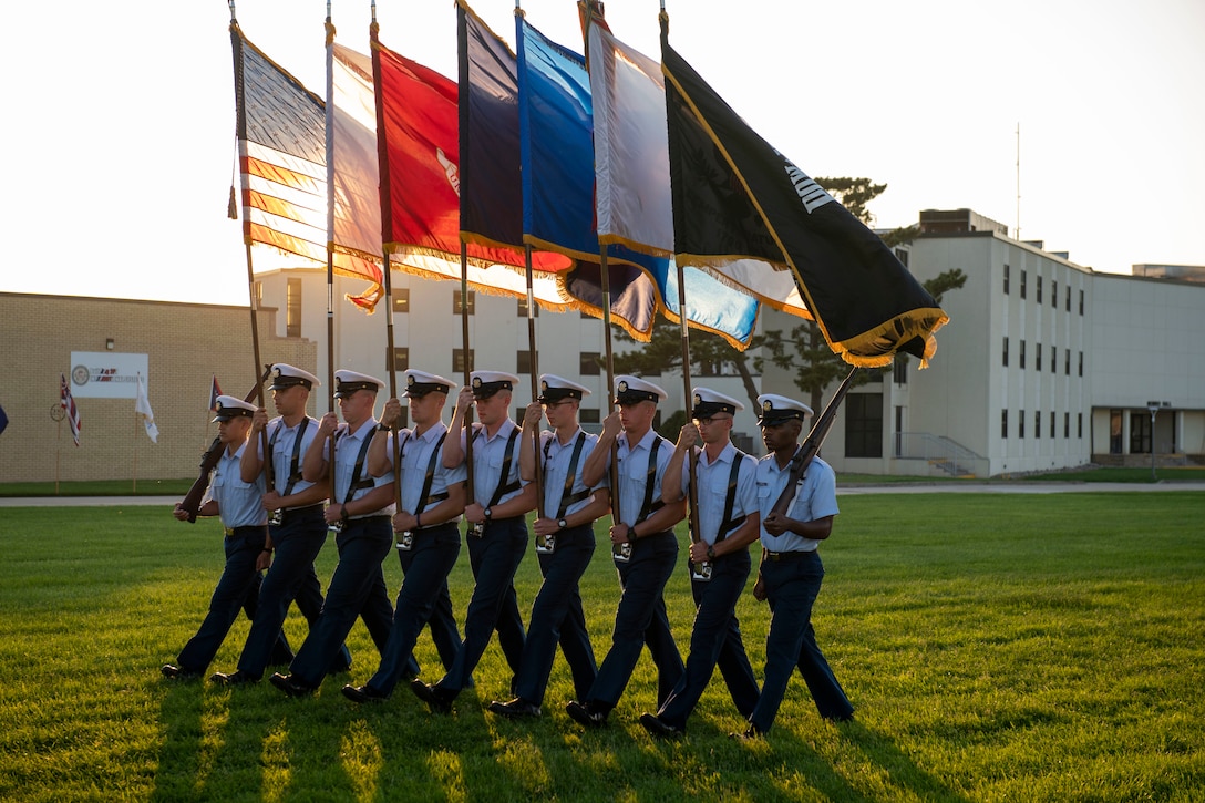 A group of Coast Guardsmen carrying flags march in formation at twilight.