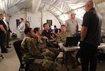photo of four USAF Airmen sitting at a table talking to two standing civilians.  Four men are standing in the back of the tent.