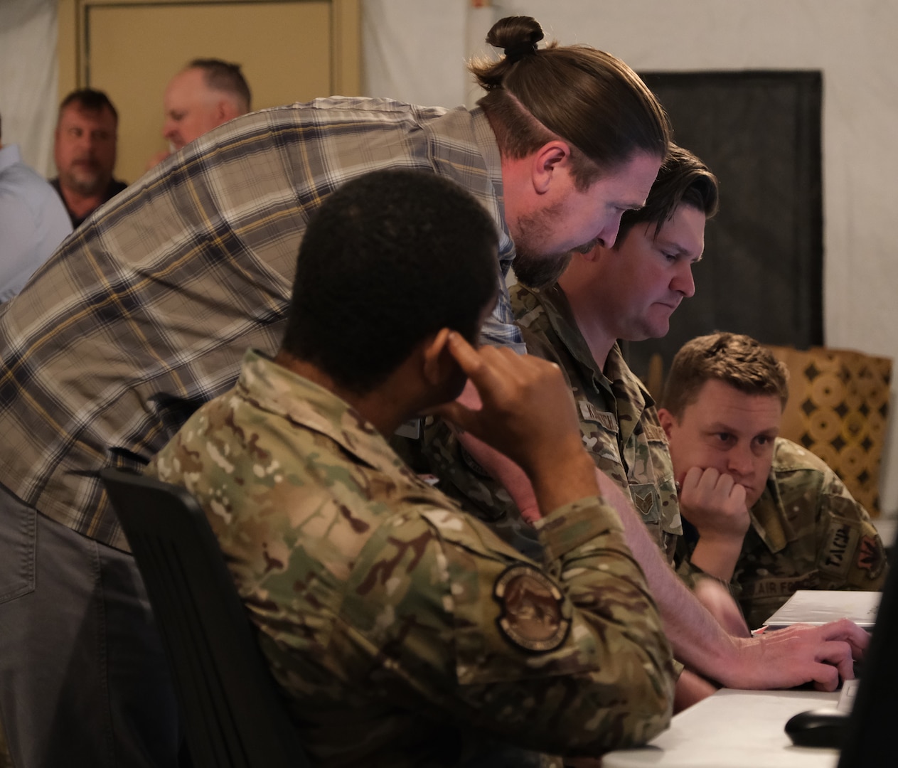 photo of three USAF Airmen sitting staring at a computer screen with a civilian standing leaning between them using a computer mouse.