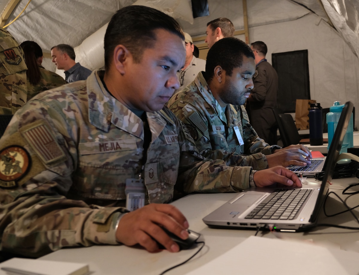 photo of two USAF Airmen sitting working on computers, with five civilian standing with their backs to the Airmen