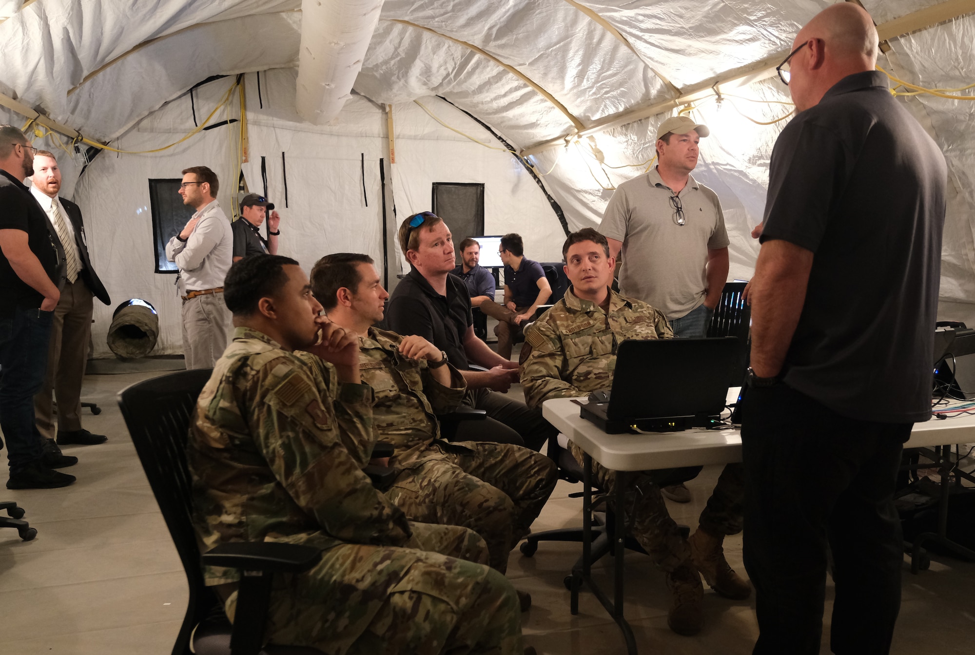 photo of four USAF Airmen sitting at a table talking to two standing civilians.  Four men are standing in the back of the tent.