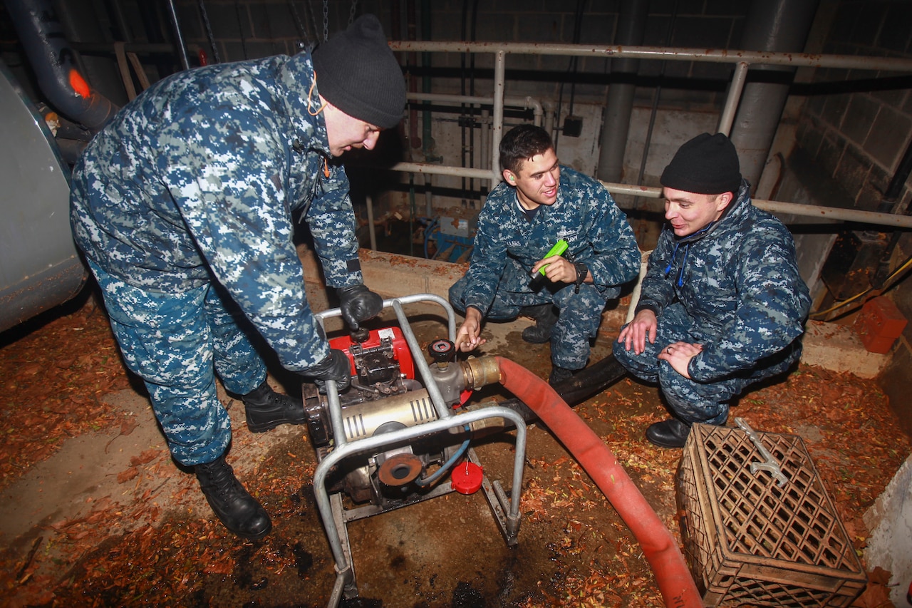 Three men operate a small portable pump in a basement.