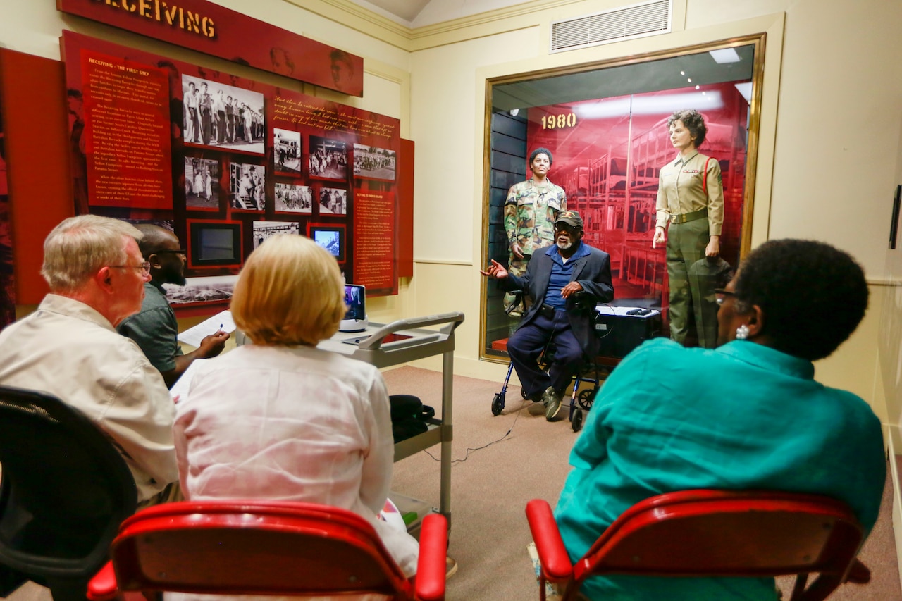 A man sitting in front of a display case talks with four other people.