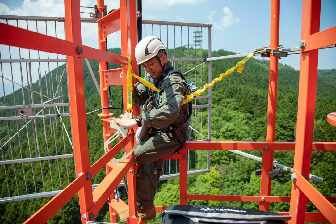 A Marine attaches a harness to a radio tower with greenery in the background.