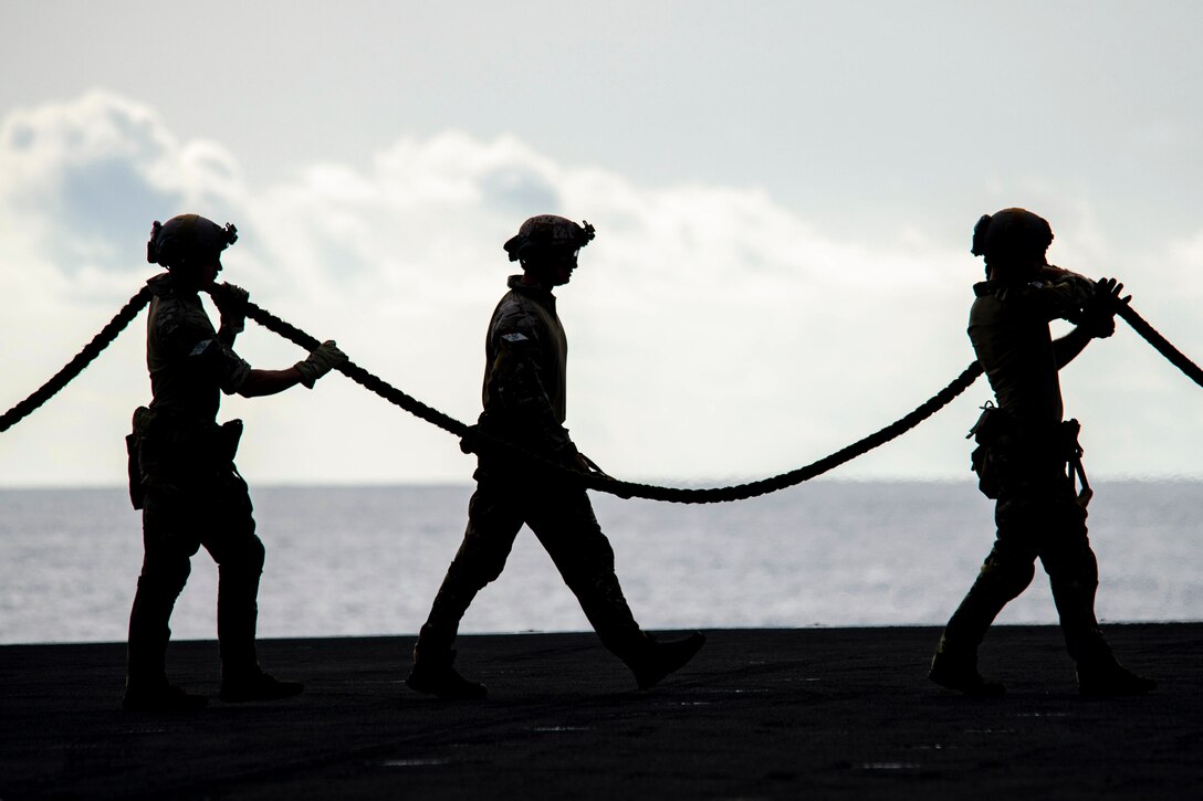 Three sailors standing in a line carry a rope aboard a ship at sea as shown in silhouette.