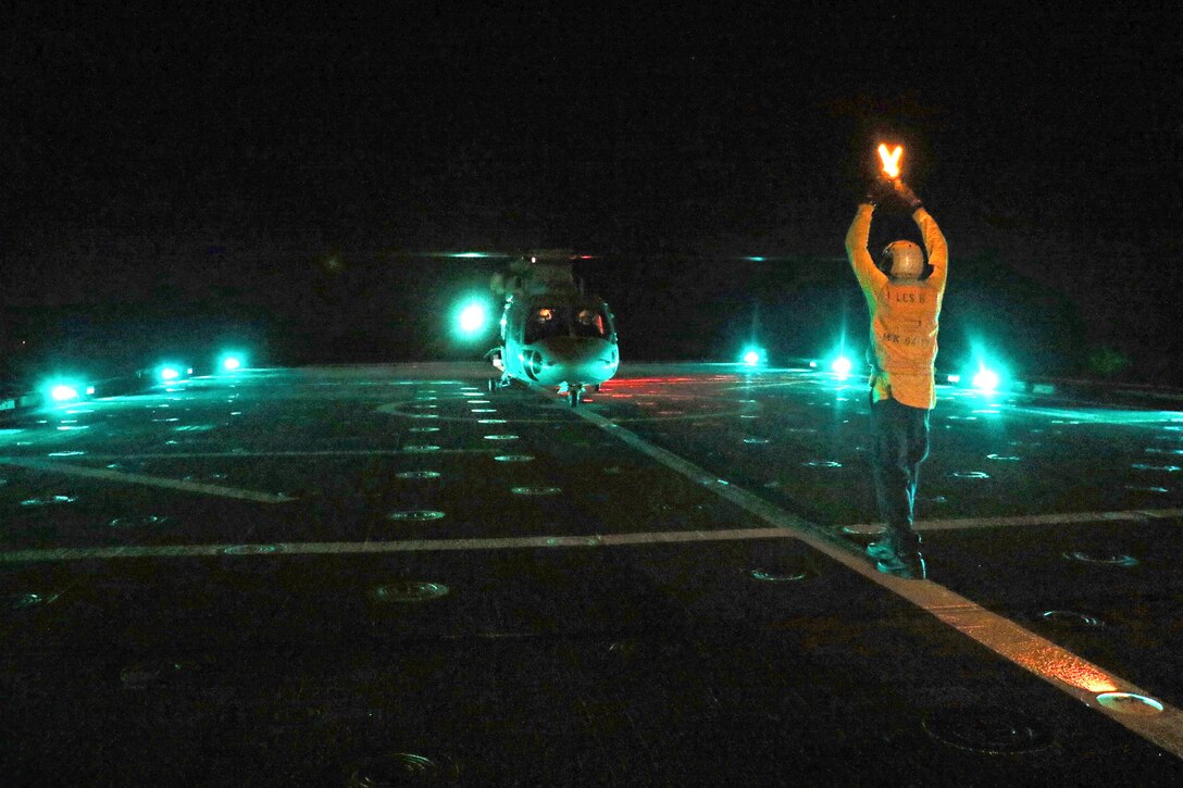 A sailor guides a helicopter on a flight deck at night.
