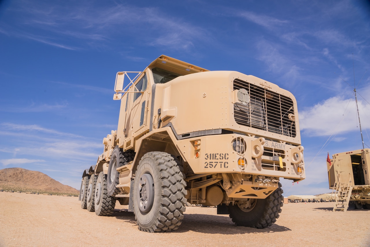 A large, dusty truck is parked on the dirt in a desert area; tents are in the background.