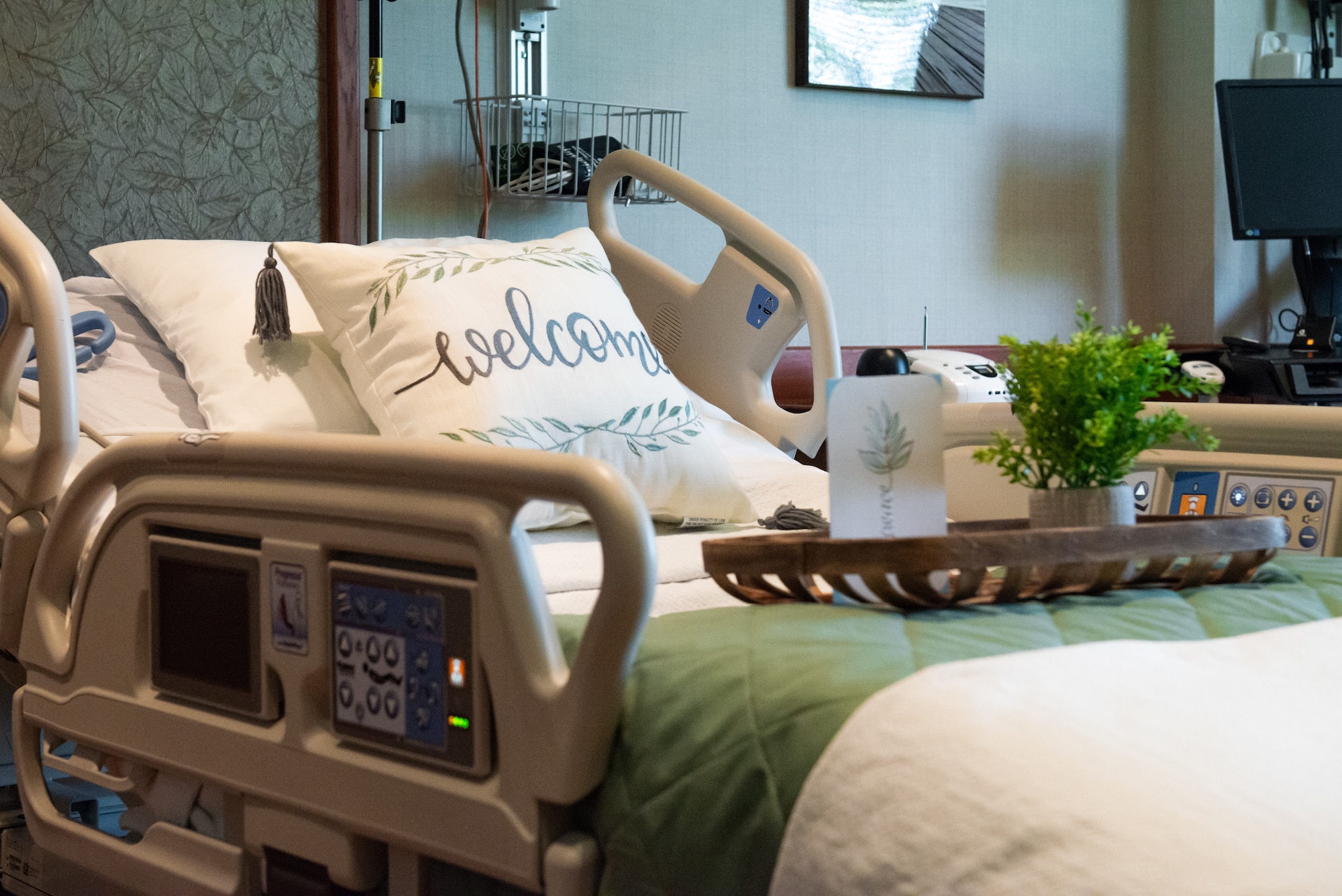 A hospital bed personalized with a white decorative pillow, a green and white blanket, and a wooden tray with a small plant on top of it.