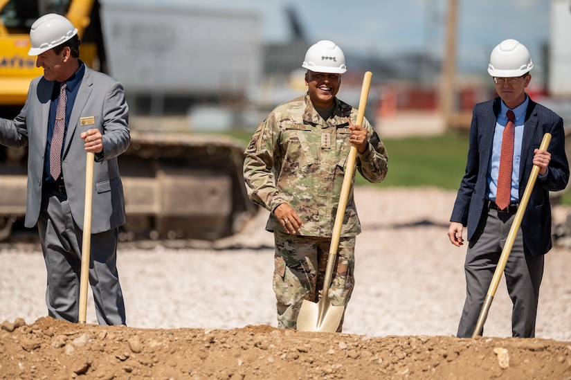 A service member and two civilians stand behind a mound of dirt and hold shovels.