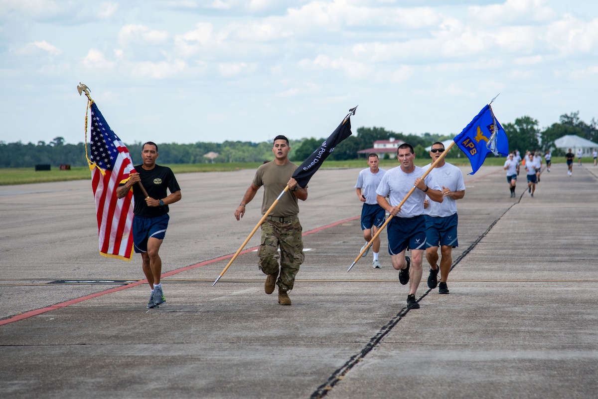 Airmen run on the flightline while carrying flags. Other Airmen are in the distant background running
