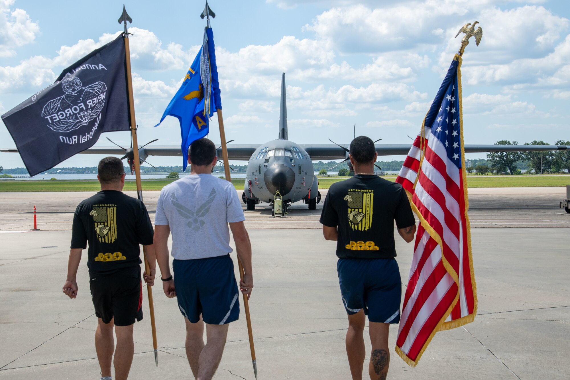 Three Airmen in PT gear walk towards the flightline bearing flags. A C-130J is in the background