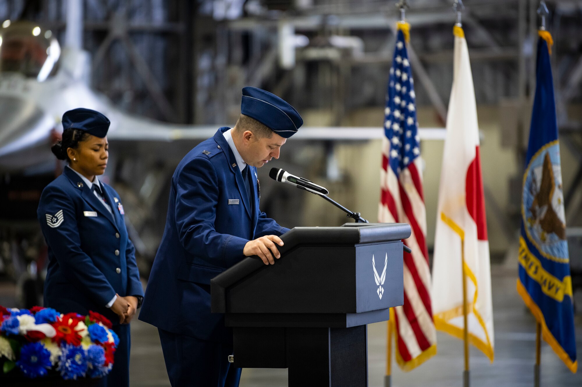 Military member in uniform talks at a podium with a F-16 Fighting Falcon in the background.