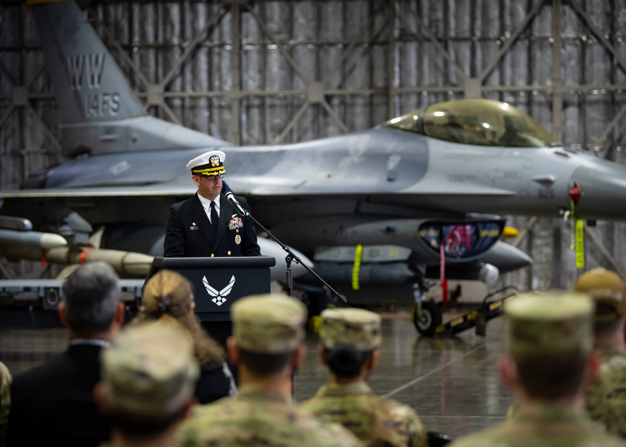 Military member in uniform talks at a podium with a F-16 Fighting Falcon in the background.