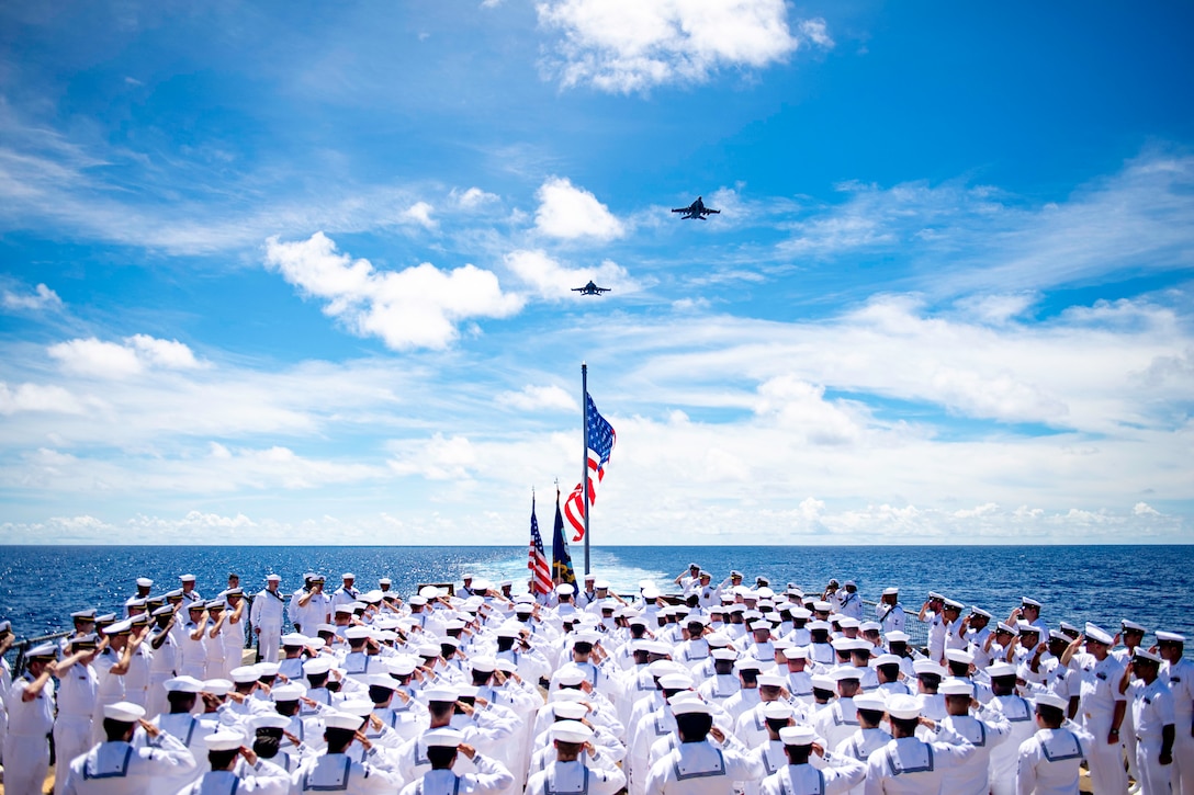 Sailors, seen from behind on a ship's deck, salute in formation as two planes fly above.